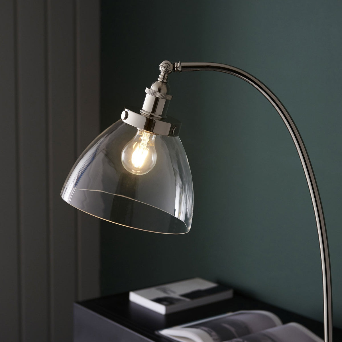 Floor lamp with a curved silver stand and clear glass shade, casting light on a dark desk.