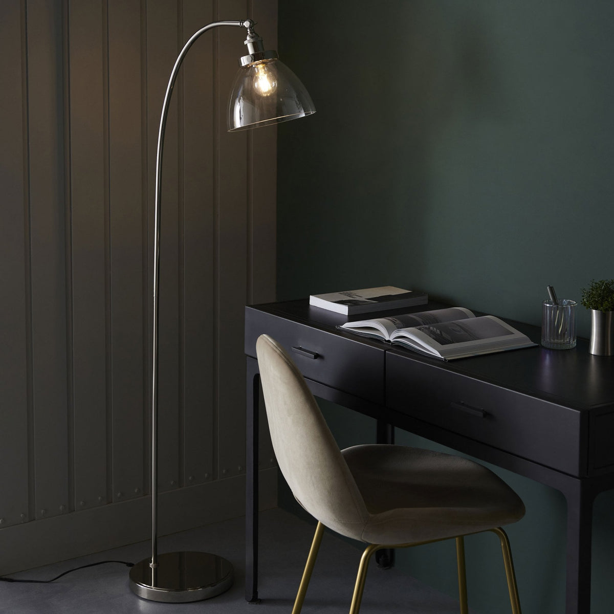 Floor lamp with a curved silver stand and clear glass shade, illuminating a dark desk with a book and chair.