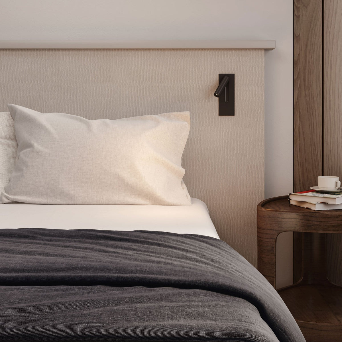 A bed with a beige headboard, white pillow, and dark grey blanket. A small lamp is on the headboard