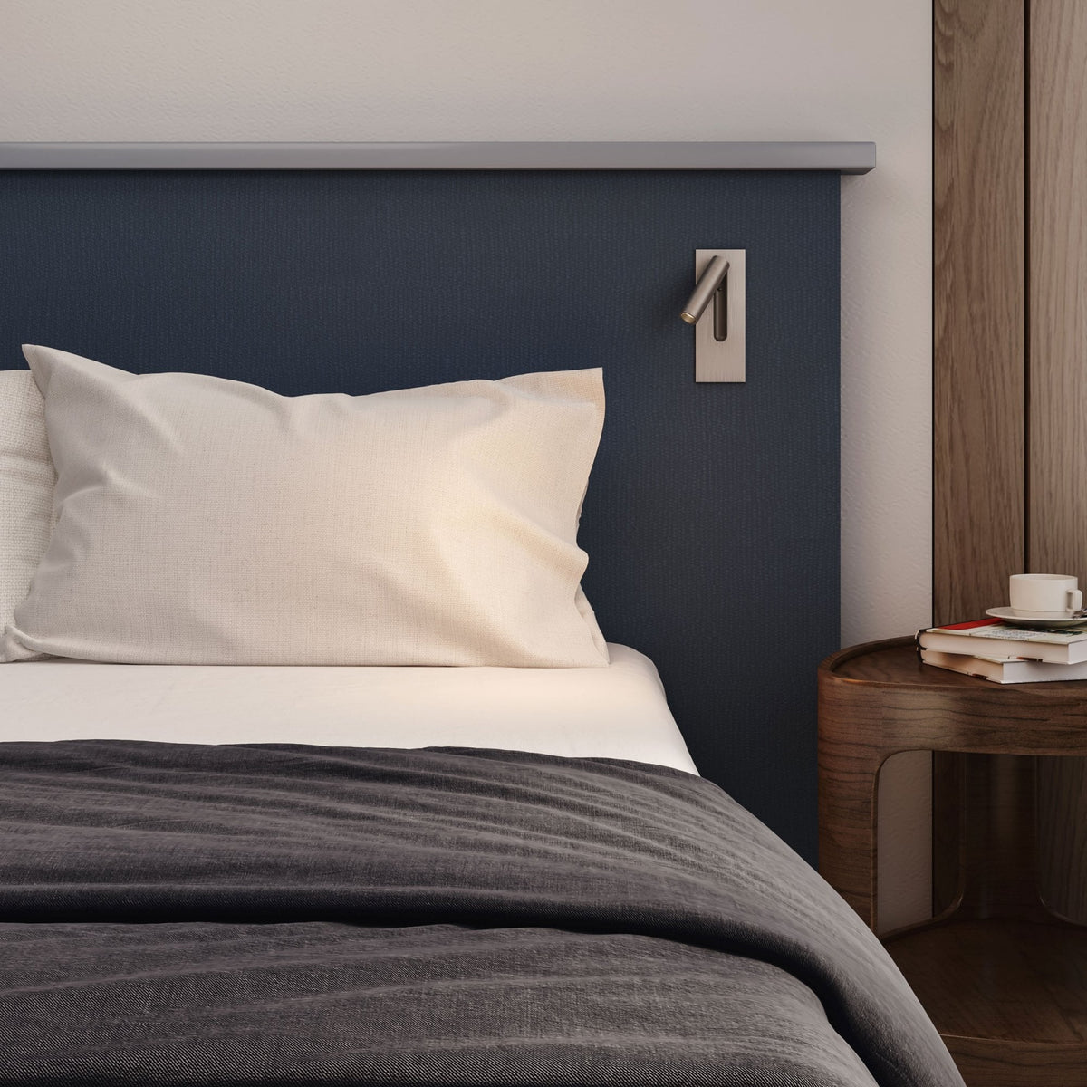 Bedroom with a dark blue headboard, white pillow, grey duvet, and a wooden nightstand with books and a cup