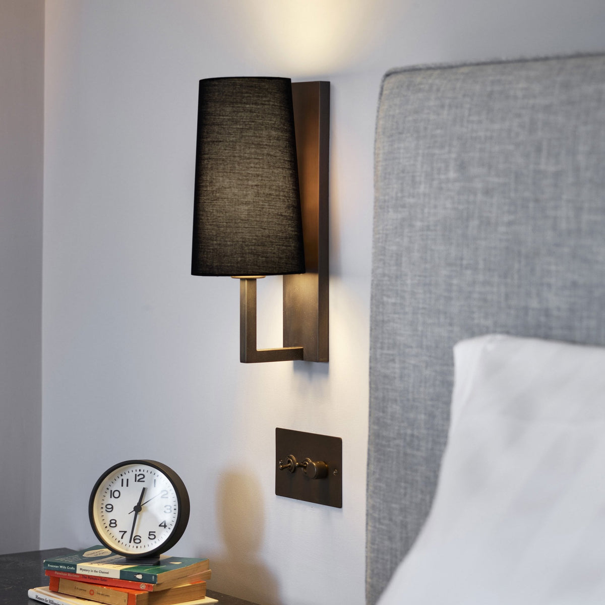 Wall-mounted lamp with black shade, light switch, alarm clock on books, and a grey headboard