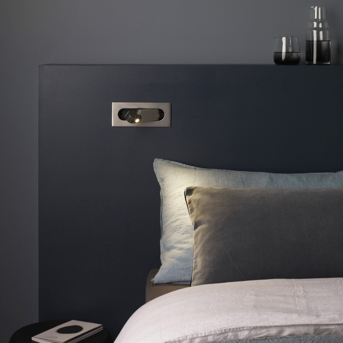 A bedroom with a dark blue wall, a light fixture, pillows, and a glass of water on a shelf