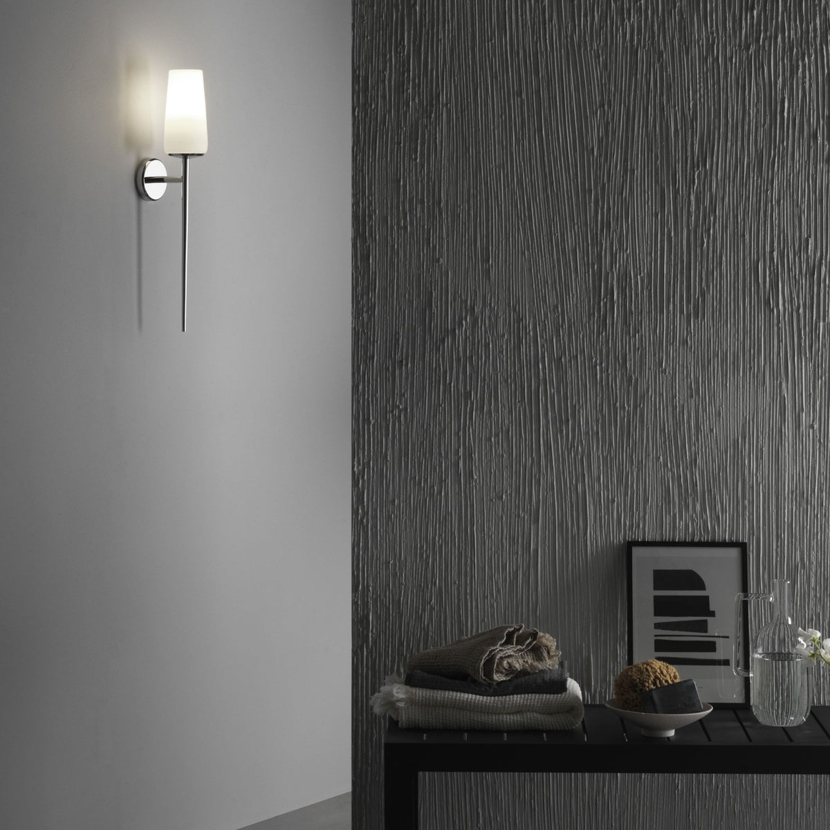 Wall-mounted light fixture with a white shade and chrome base, illuminating a textured gray wall
