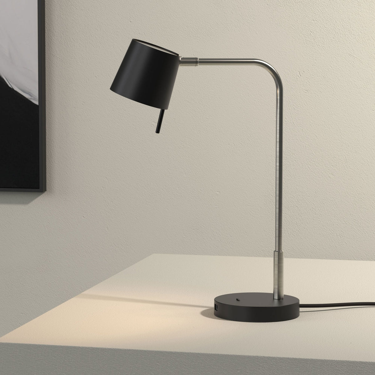 Black desk lamp with a brushed steel arm and base on a white surface, with a black framed picture in the background