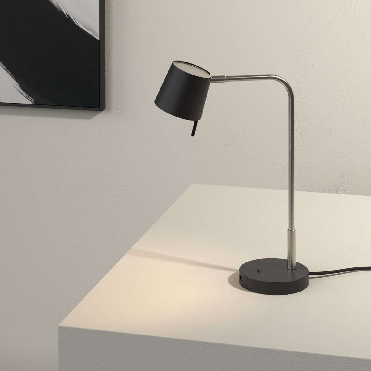 Black desk lamp with a brushed steel arm and base, illuminating a white surface next to a black and white painting