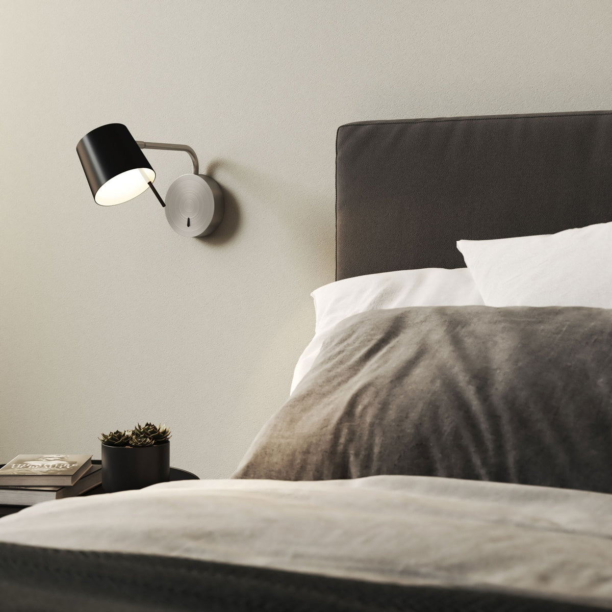 Black wall lamp with a brushed metal base and arm, illuminating a bedside table with books and a plant
