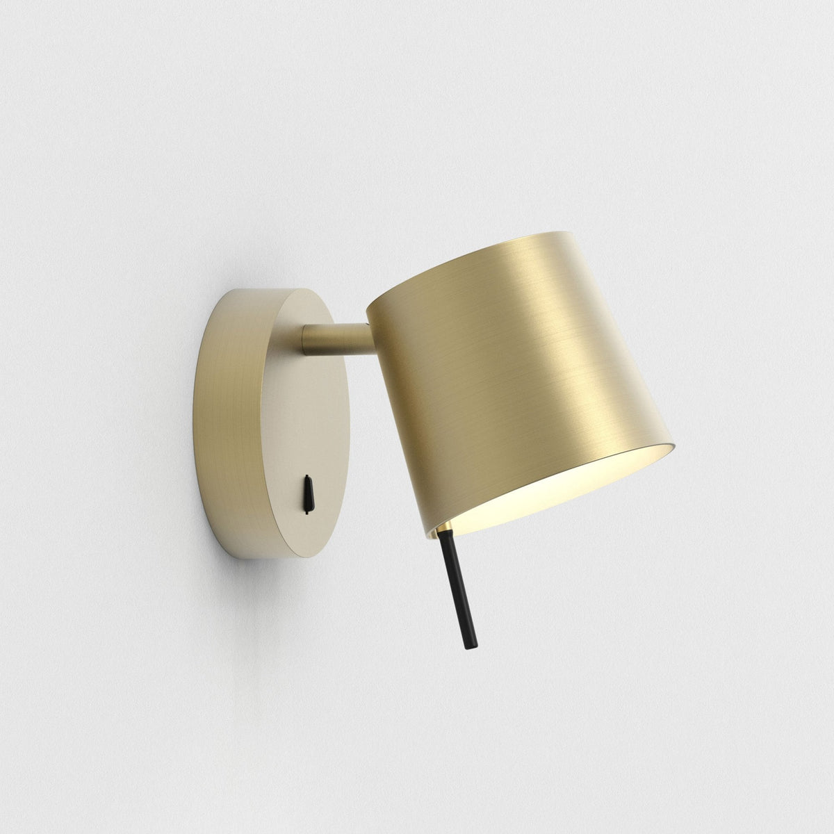 Brushed brass wall light with a cylindrical shade angled downwards, emitting a warm glow