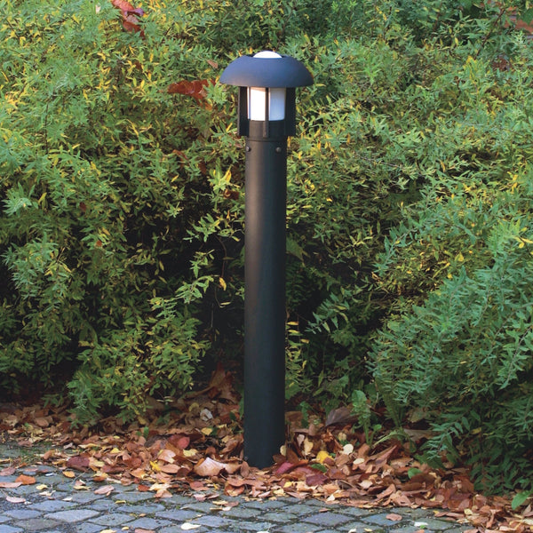 A black bollard light stands in a garden with green bushes and autumn leaves.