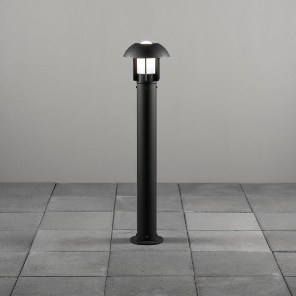 A modern black bollard light illuminated on a grey tiled patio.