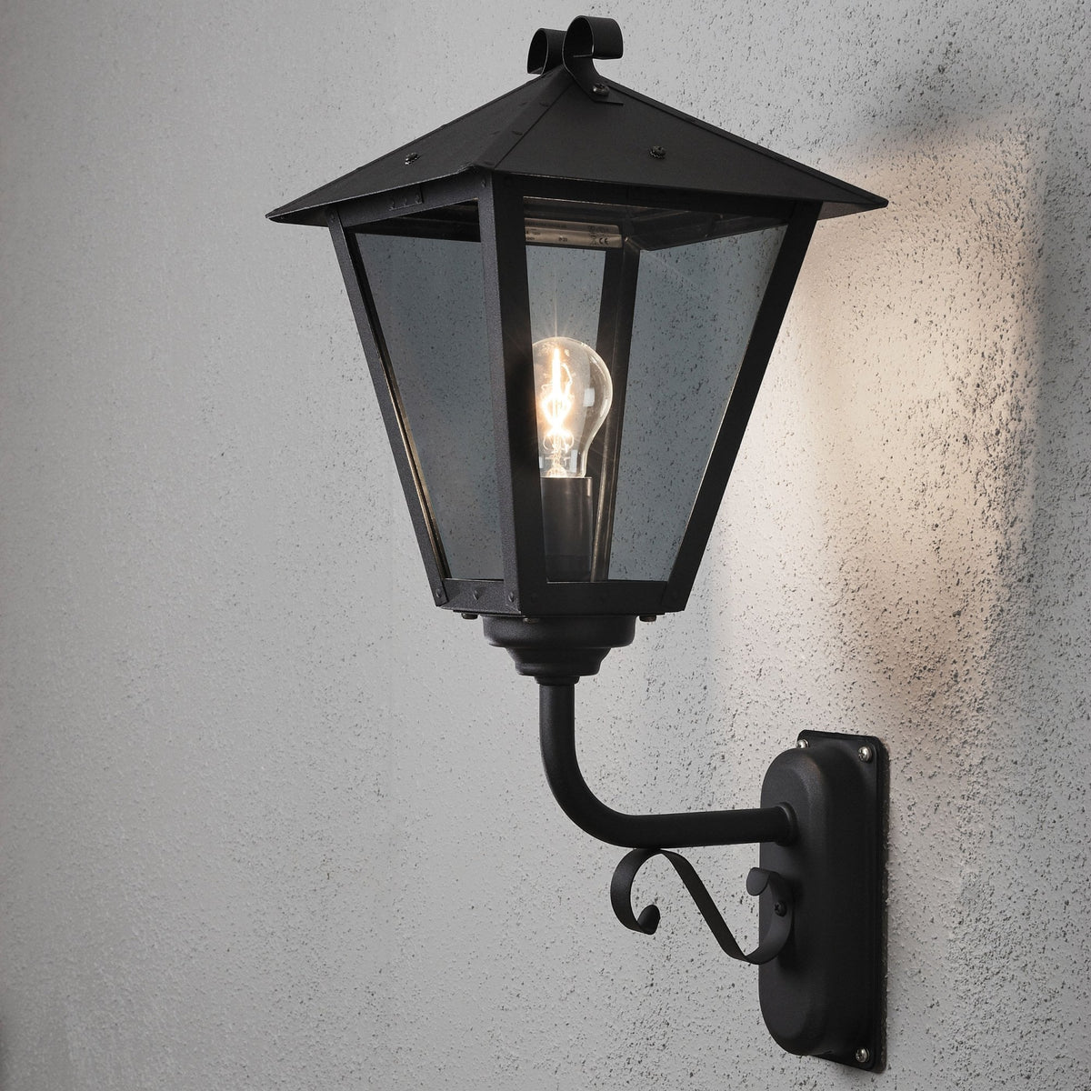A black outdoor wall light with clear glass and a visible bulb, casting light on a wall.