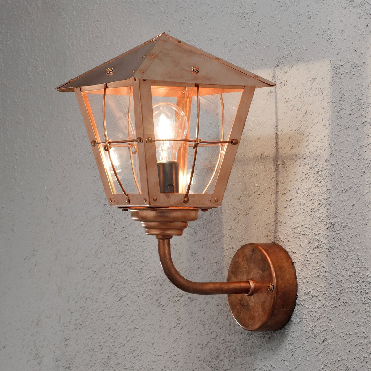 A close-up of a bronze outdoor wall light with a clear glass shade, seen from a slight angle.