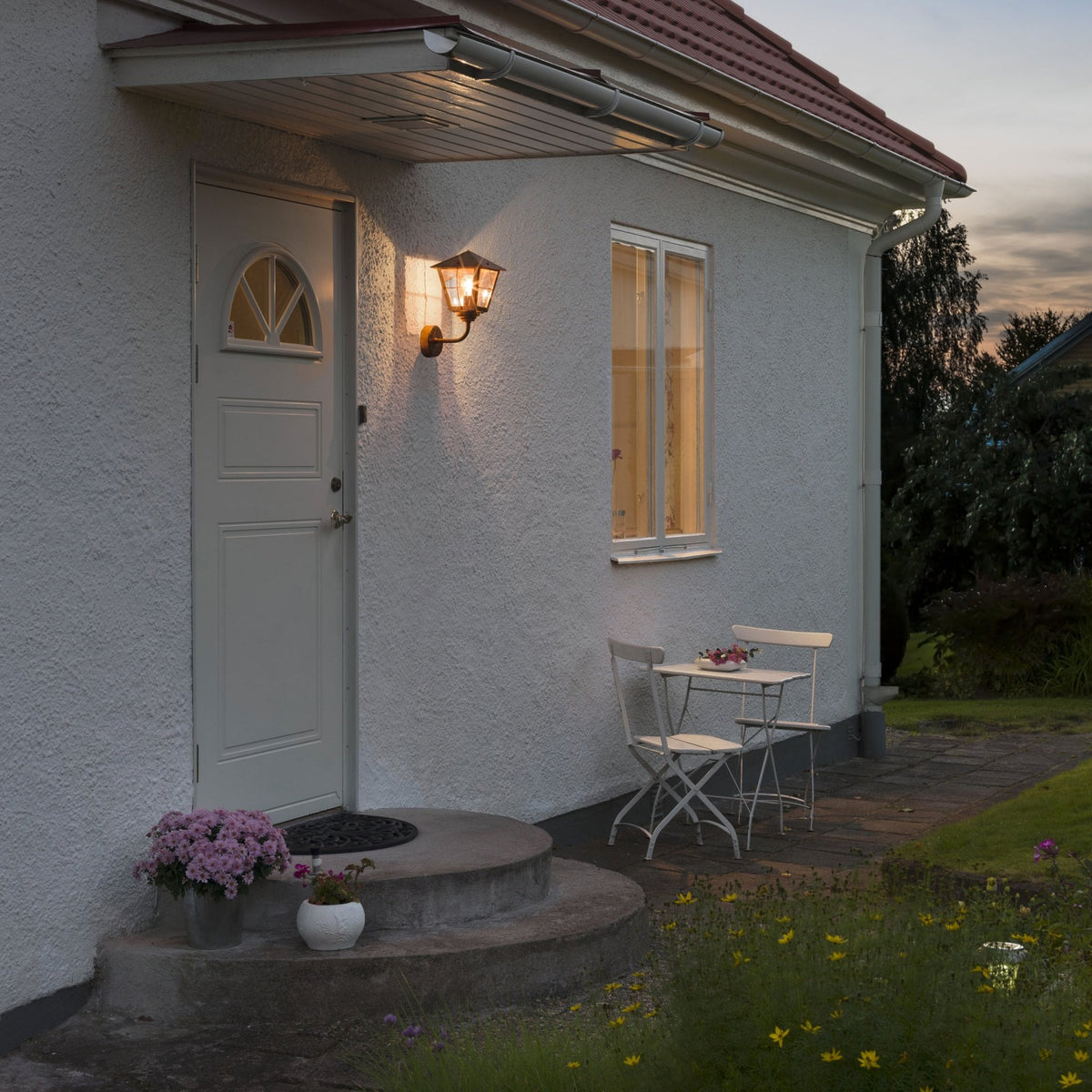 An outdoor wall light illuminating the entrance of a house at dusk.
