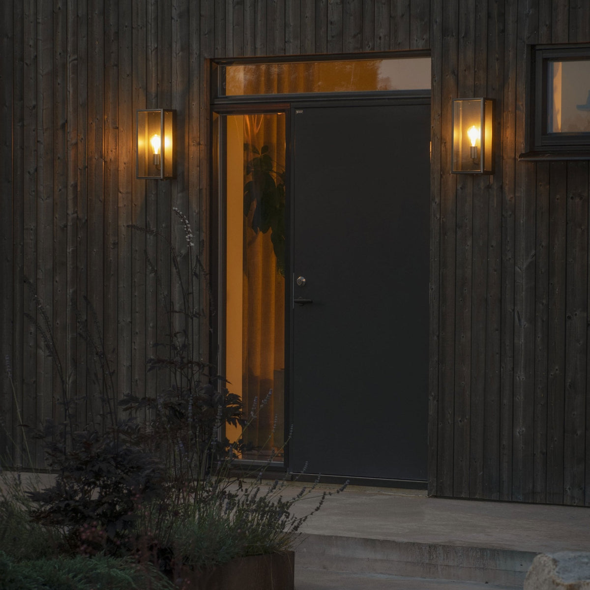 Two outdoor wall lights illuminating a dark grey front door on a modern wooden house.