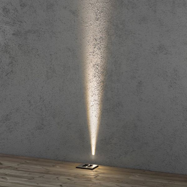 A slim, warm light beam from a ground light illuminates a concrete wall and wooden floor.