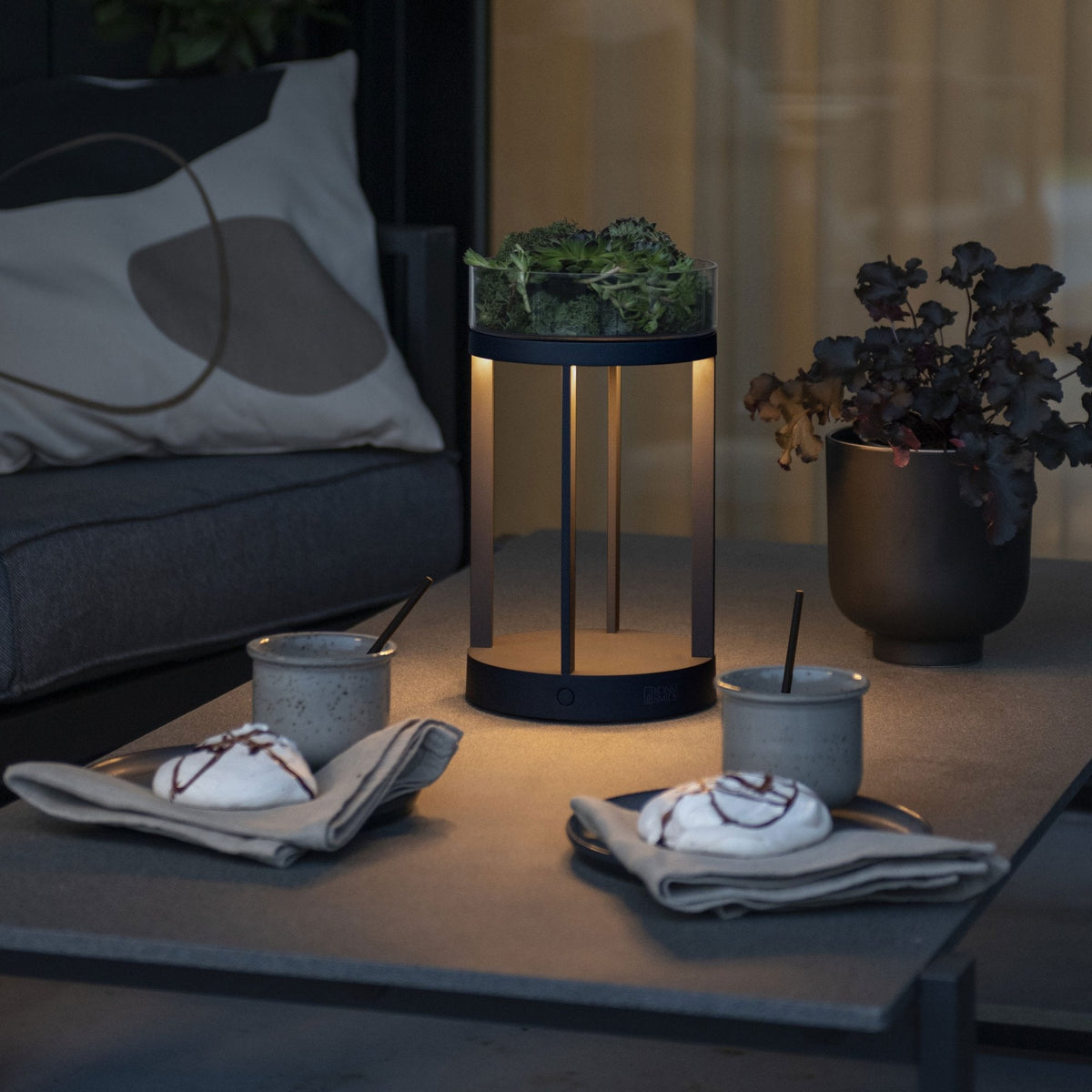 A modern outdoor lantern illuminating a table with desserts and drinks at dusk.