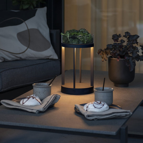 A modern outdoor lantern illuminating a table with desserts and drinks at dusk.