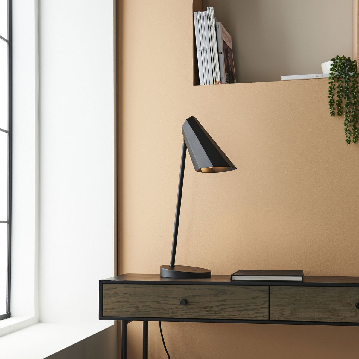 Black desk lamp on a dark wooden console table next to a window.