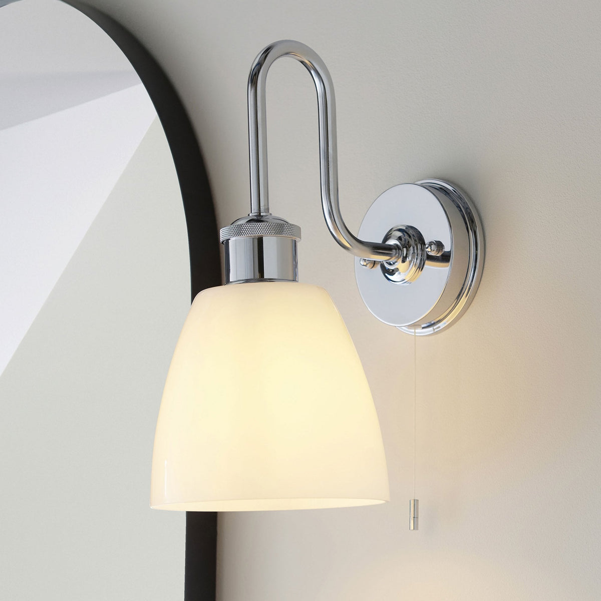 Chrome wall light with a curved arm and a white bell-shaped shade, next to a black-framed mirror.