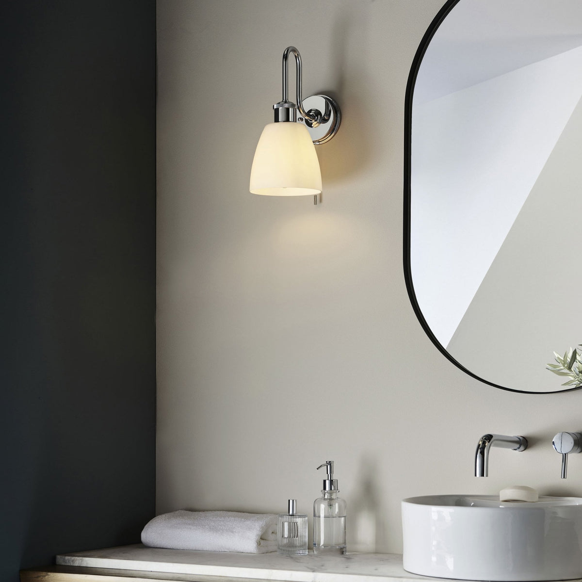 Chrome wall light with a white shade illuminating a modern bathroom vanity with a white sink and mirror.
