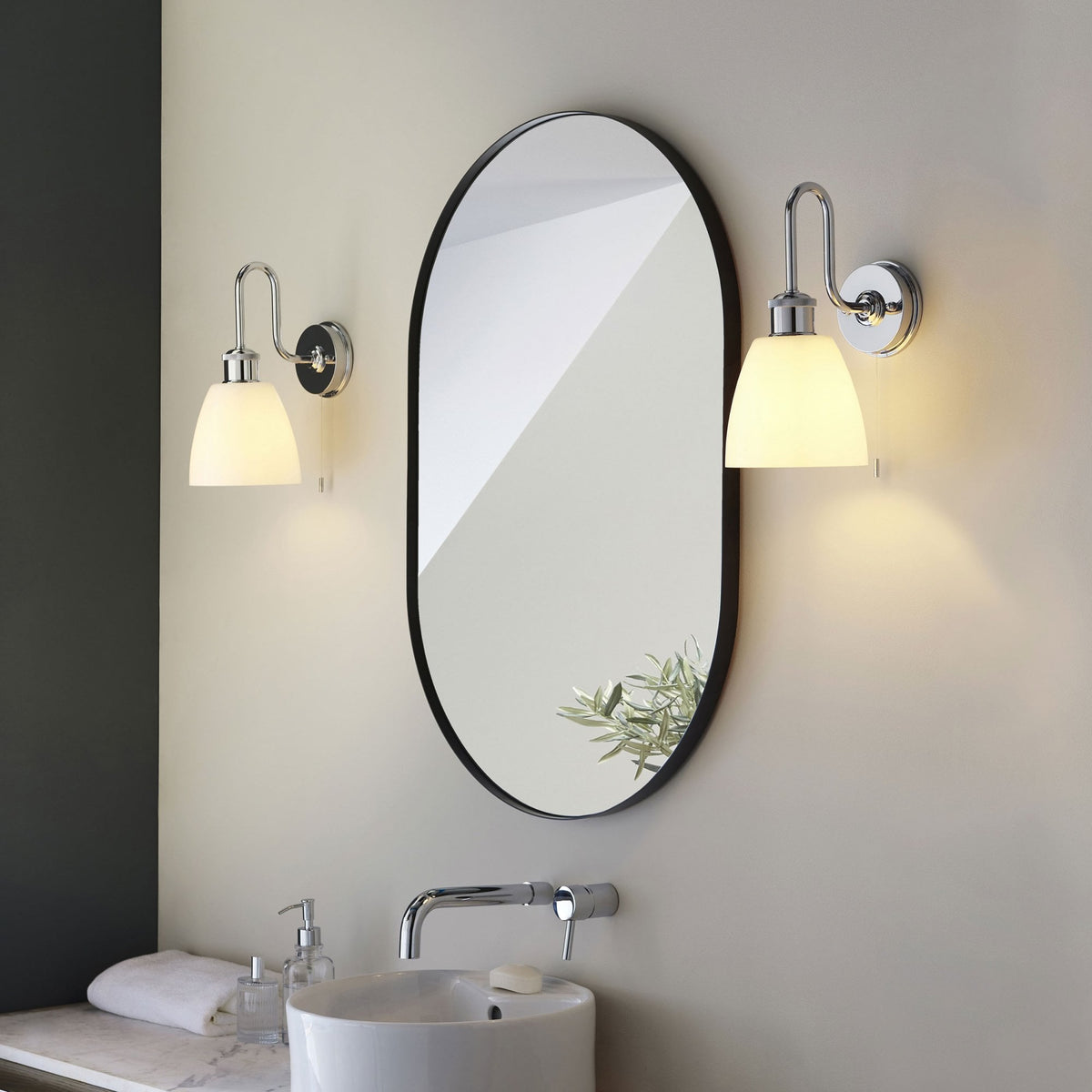 Two chrome wall lights with white shades flanking an oval mirror above a modern bathroom sink.