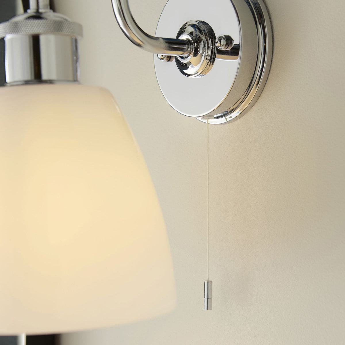 Close-up of a chrome wall light with a white shade and a silver pull cord switch against a light wall.