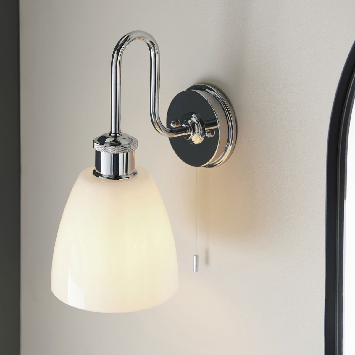 Chrome wall light with a curved arm and a white bell-shaped shade, casting a soft light on a wall.