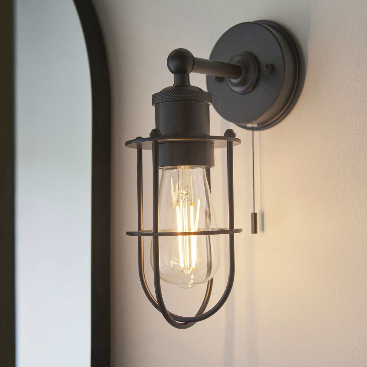 Black caged wall light with a pull cord switch and exposed bulb, casting a warm glow on a light wall next to a mirror.
