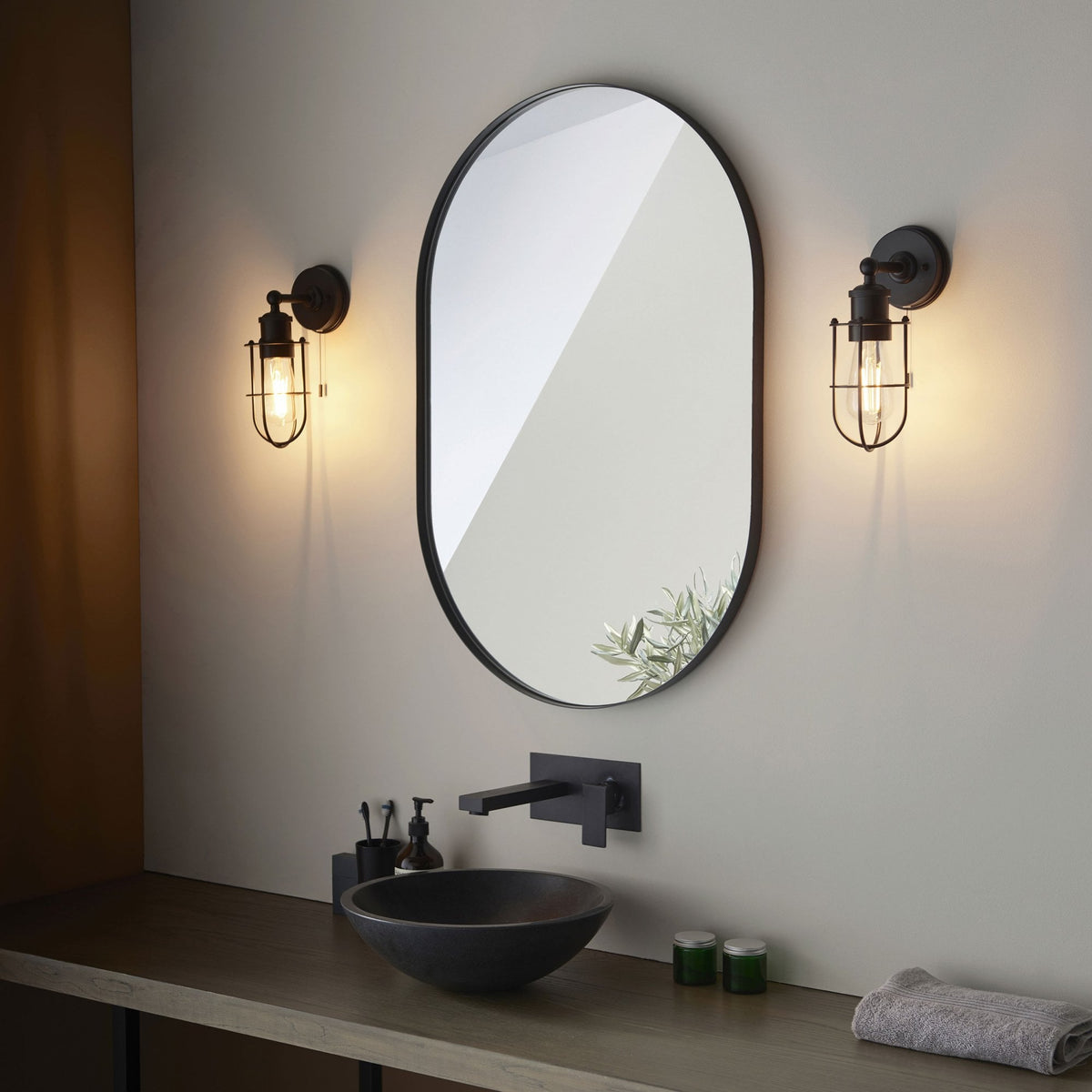 Two black caged wall lights flank an oval mirror above a modern bathroom vanity with a black basin and faucet.