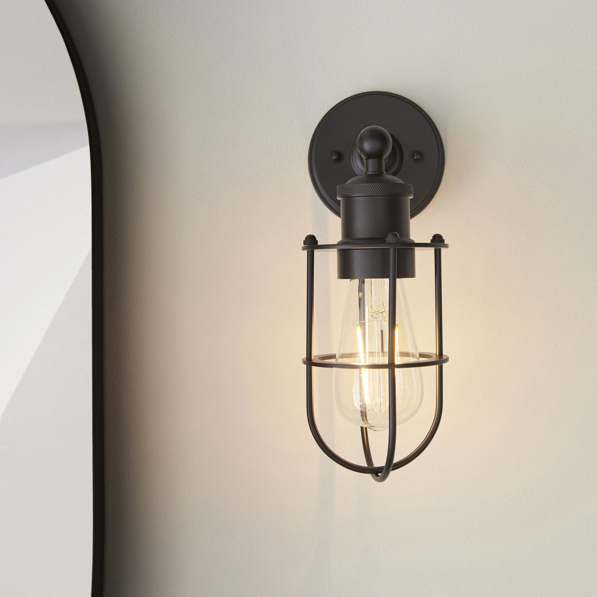 Black caged wall light with an exposed bulb, illuminating a light-colored wall next to a curved mirror.