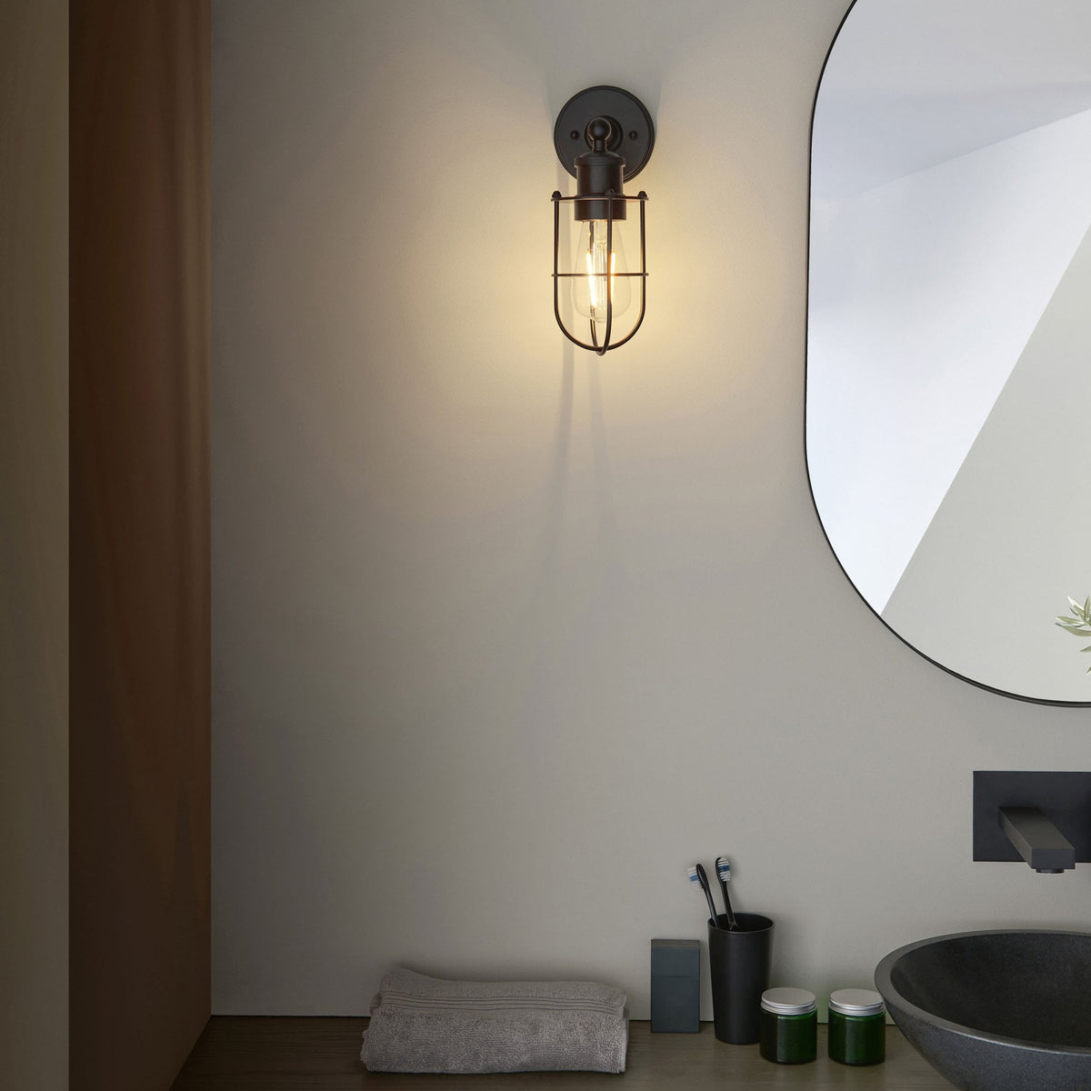 Black caged wall light with an exposed bulb, casting light on a modern bathroom vanity with a black basin and mirror.