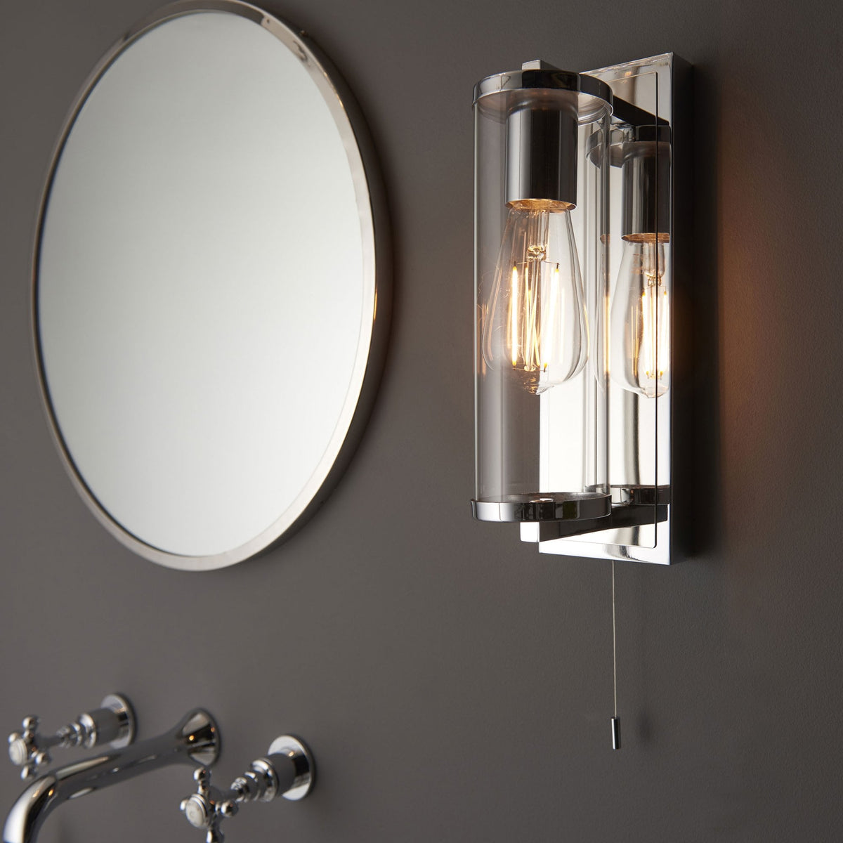 Chrome wall light with two exposed bulbs and a pull cord, next to an oval mirror and sink taps.