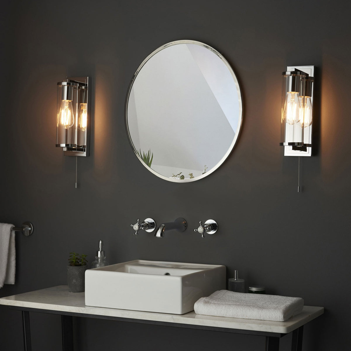 Two chrome wall lights with exposed bulbs flank an oval mirror above a modern sink and dark wall.