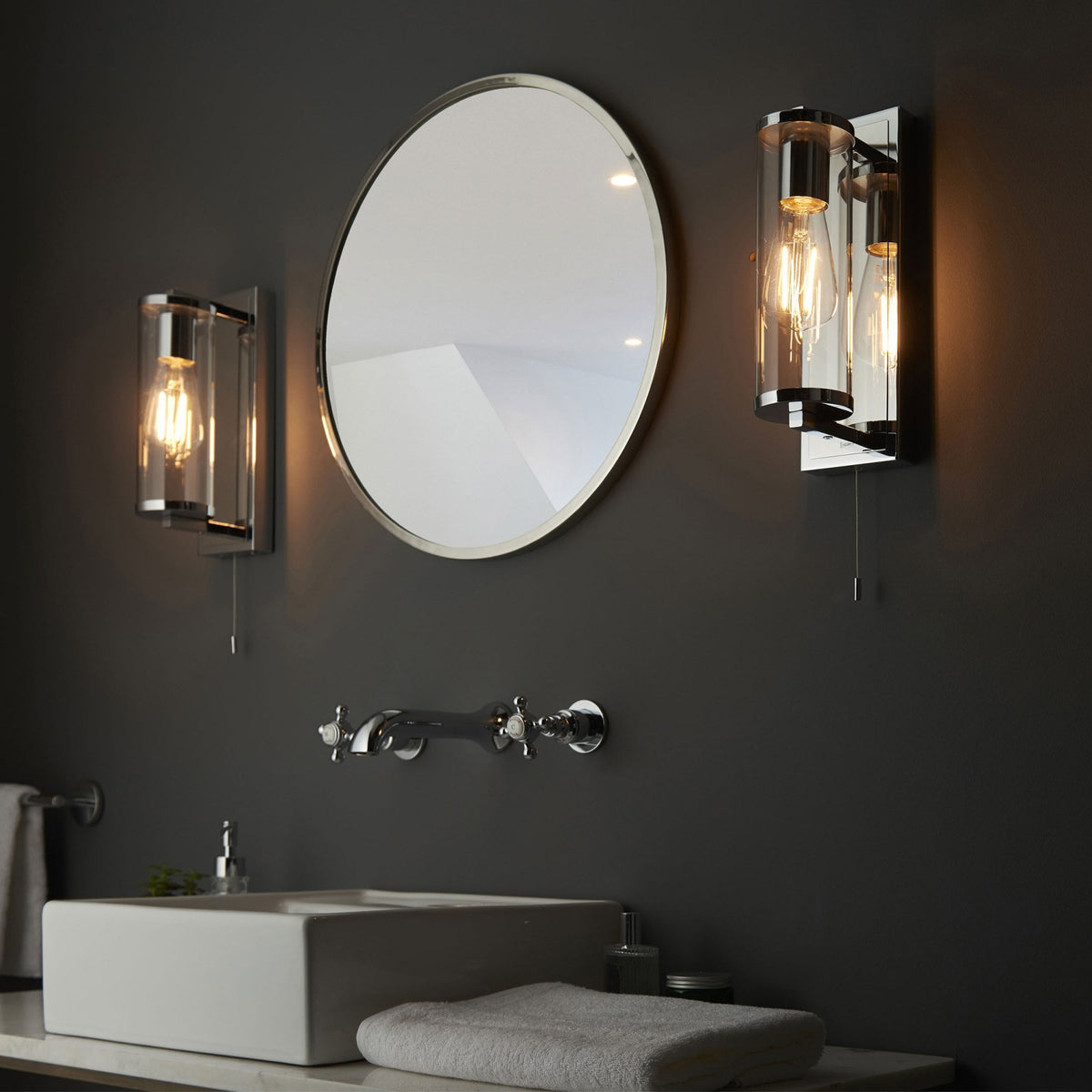 Two chrome wall lights with exposed bulbs and pull cords, framing an oval mirror above a sink.