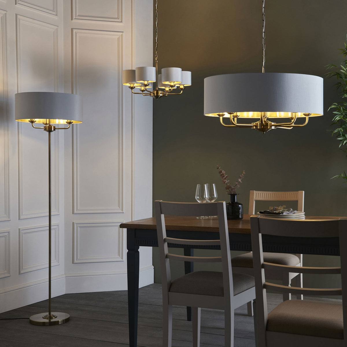 A pendant light and a floor lamp, both with white drum shades and brass interiors, illuminate a dining room.