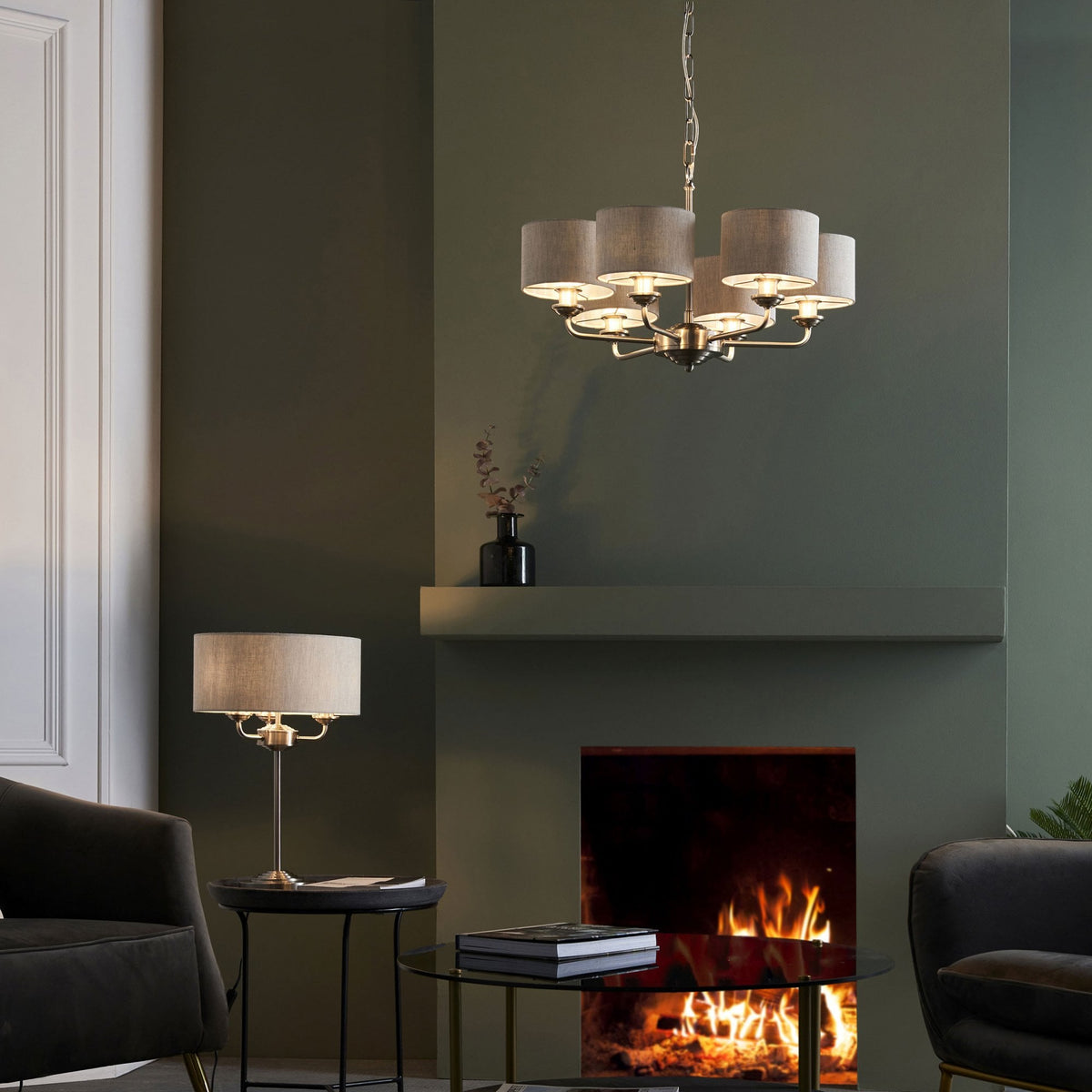 A five-light chandelier and matching table lamp illuminate a living room with a lit fireplace.