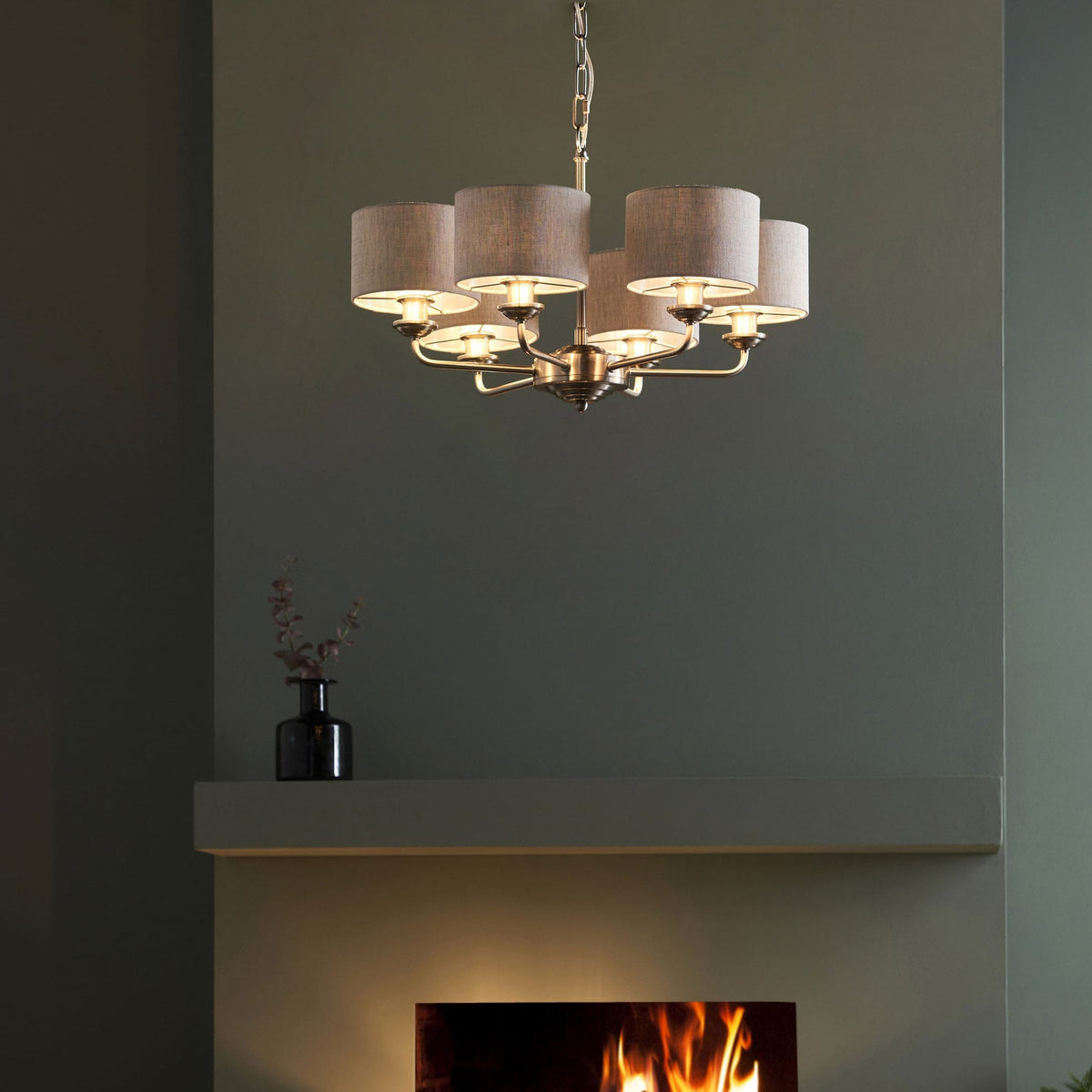 A five-light chandelier with grey fabric shades hangs above a fireplace with a warm glow.
