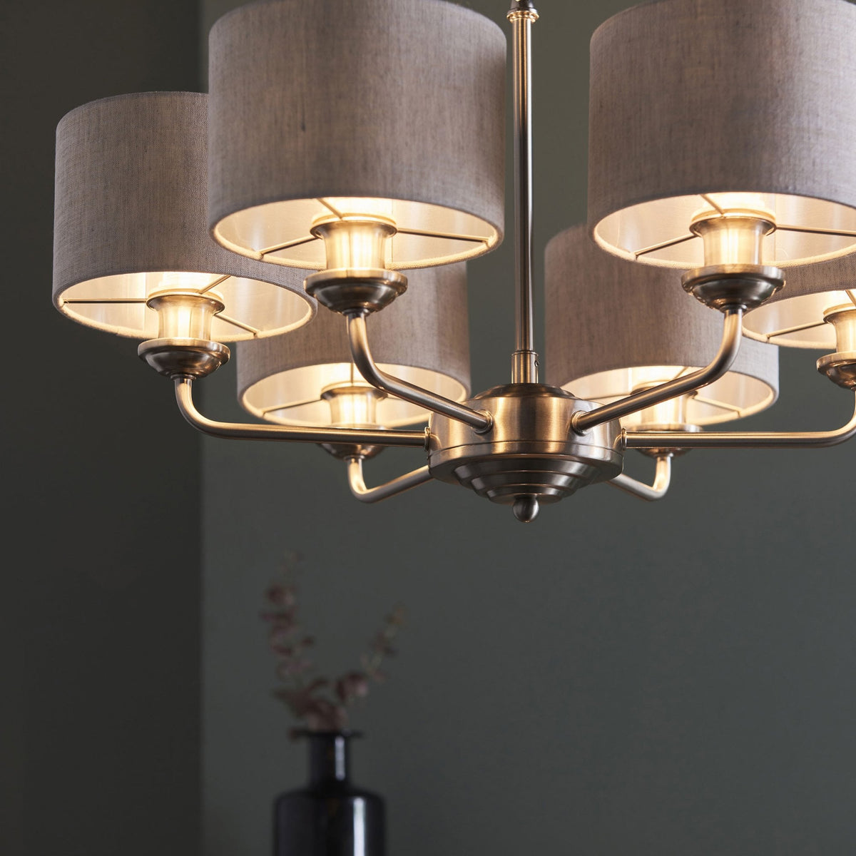 Close-up of a five-light chandelier with grey fabric shades, illuminating a dark green wall.