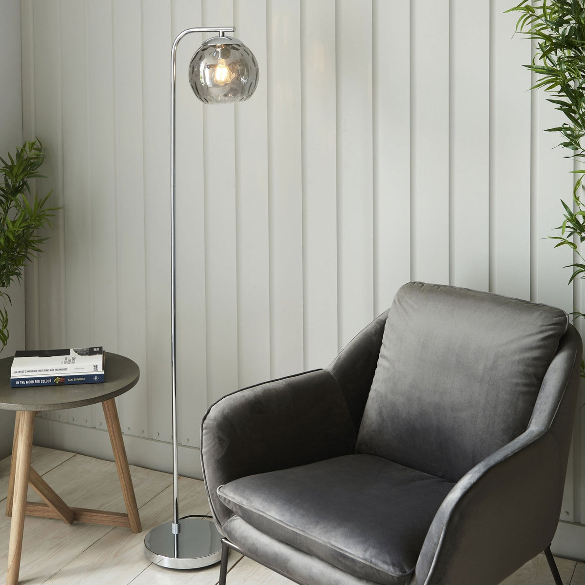 Chrome floor lamp with a smoked glass shade positioned beside a grey velvet armchair and a small side table.