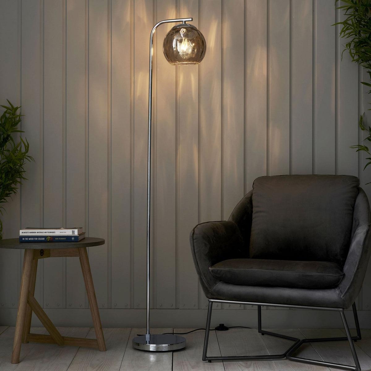 Chrome floor lamp with a glowing smoked glass shade illuminating a grey armchair and a wooden side table.