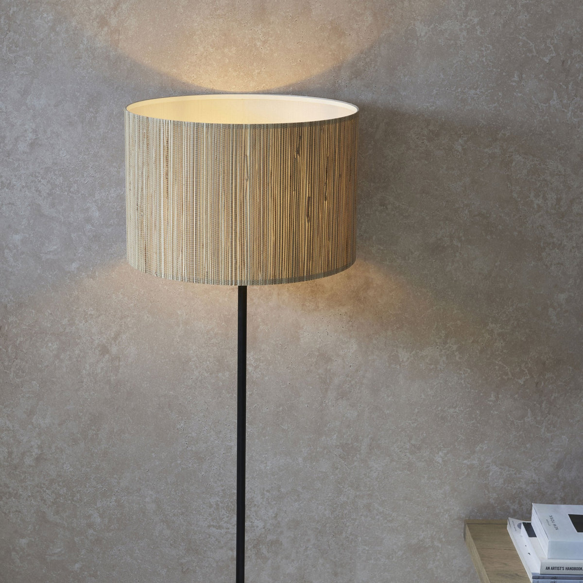 Close-up of a floor lamp's natural woven shade, casting a warm glow on a textured grey wall.
