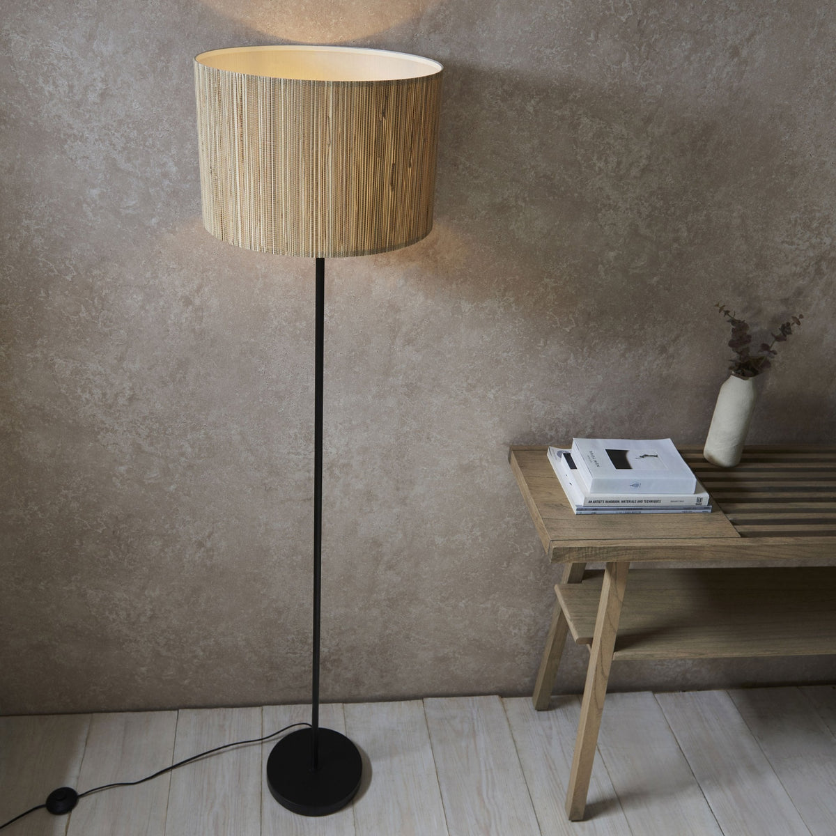 A floor lamp with a natural woven shade stands beside a light wooden table with books and a vase.