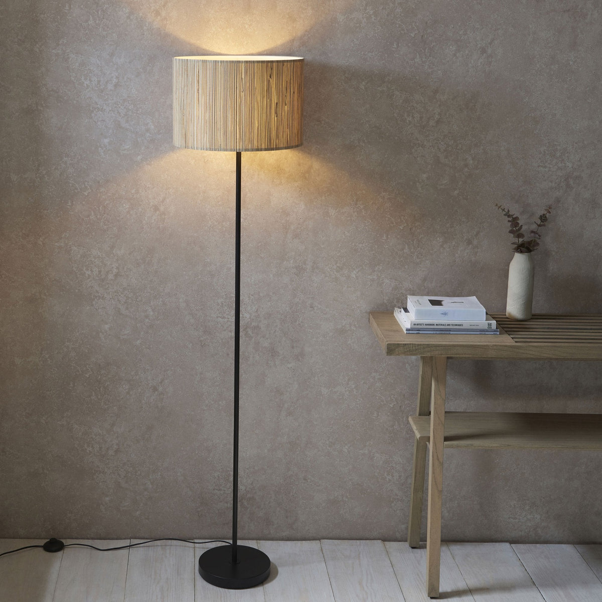 A floor lamp with a natural woven shade illuminates a room with a light wooden table and a white vase.