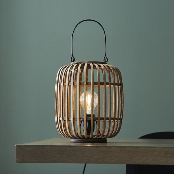 A bamboo cage table lamp on a light wooden table against a dark wall.