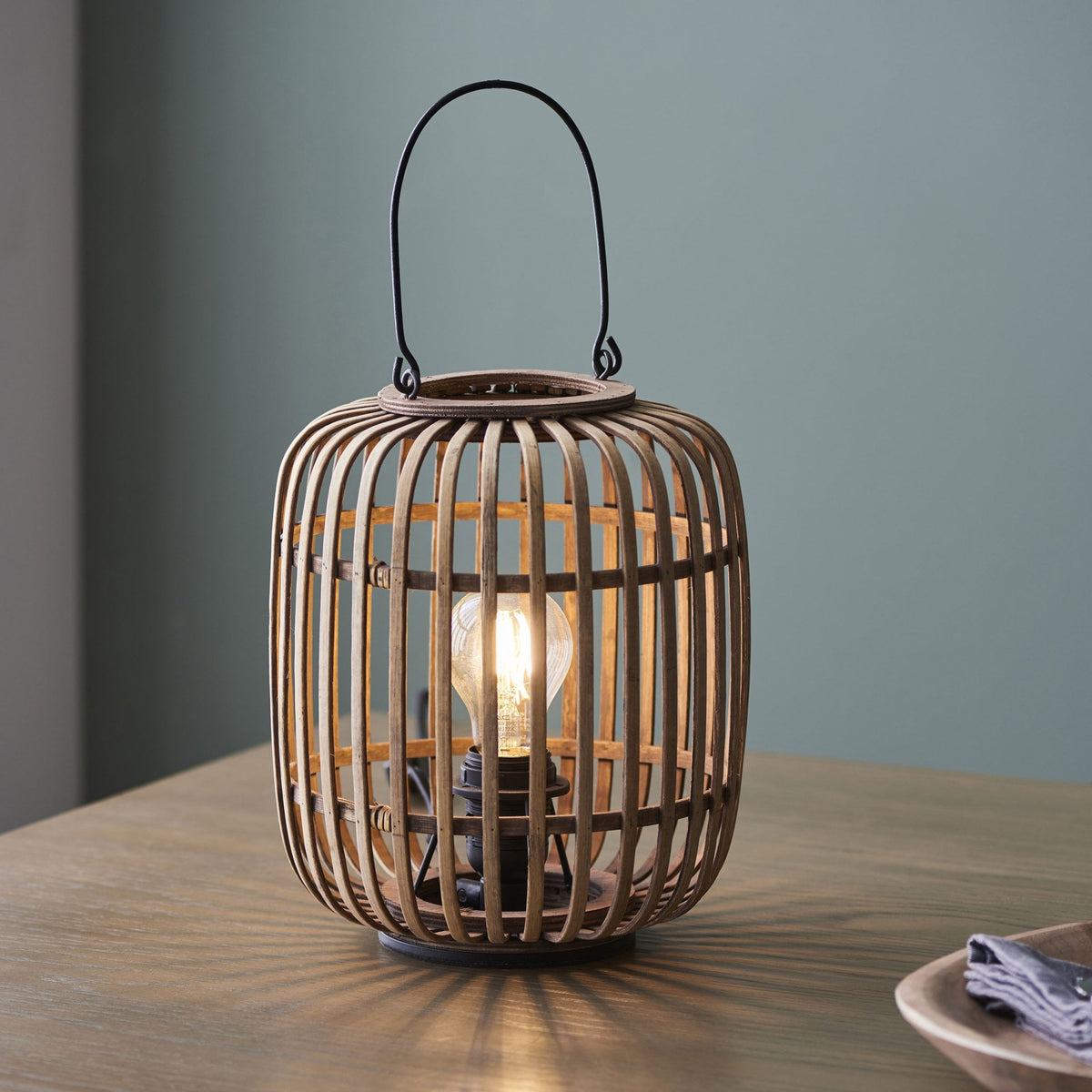 A bamboo cage table lamp casting a glow on a light wooden table.