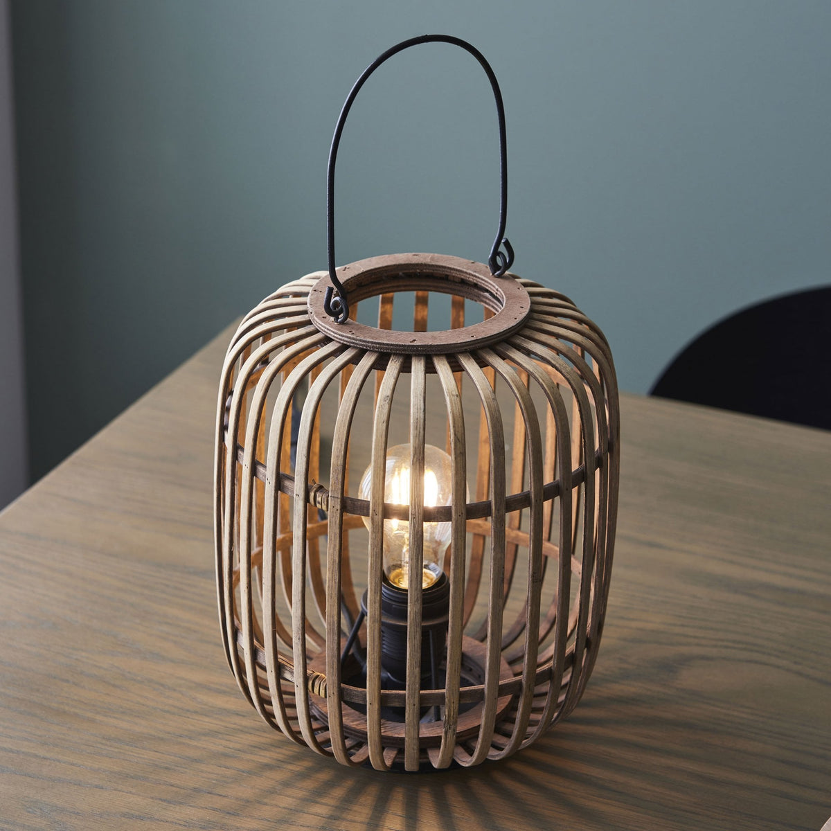 A bamboo cage table lamp with the light on, on a wooden surface.