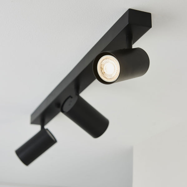 A close-up of a black track light with three adjustable spotlights, one illuminated, on a white ceiling.