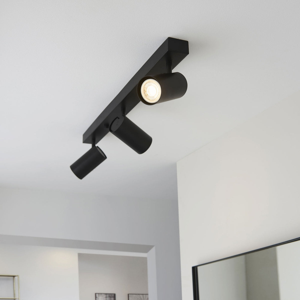 A black track light with three spotlights illuminates a contemporary hallway with white walls.