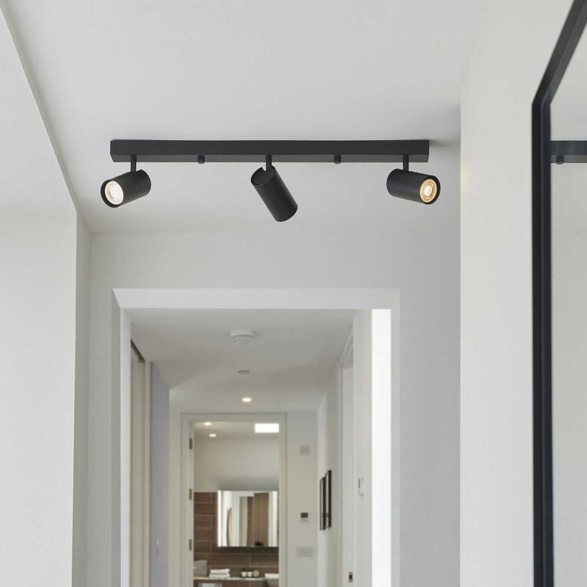 A black track light with three spotlights illuminates a modern hallway with white walls and a mirror.