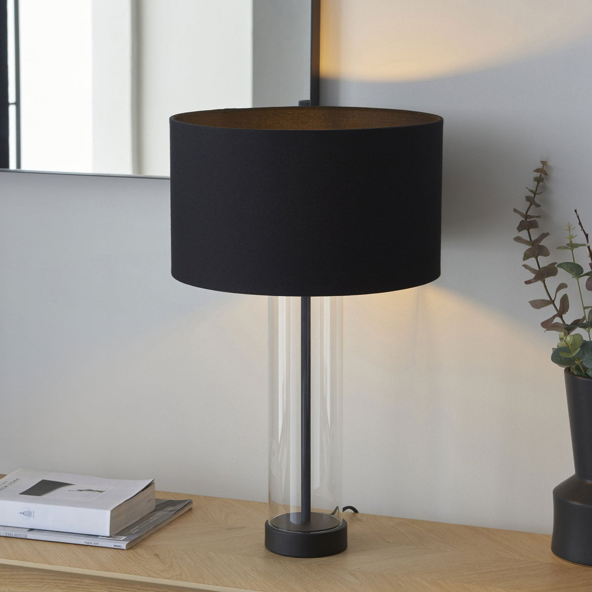 A black table lamp with a clear glass base sits on a wooden surface next to a dark vase with greenery.