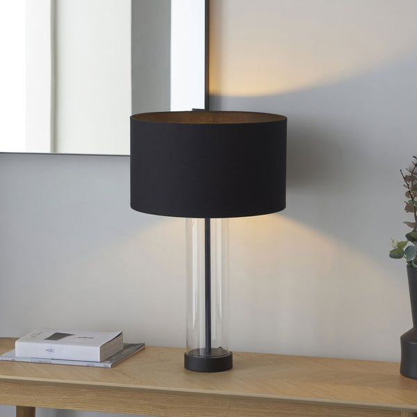 A black shade table lamp with a clear glass base, illuminated, on a wooden console table.